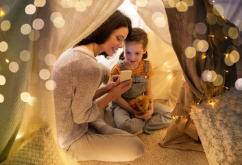 Sticker - family, hygge and technology concept - happy mother and little daughter with smartphone in kids tent at night at home