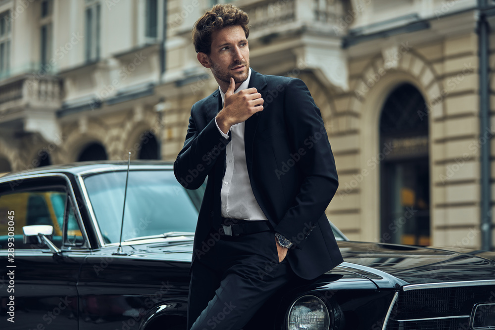 Young handsome man with black classic car wearing black suit - obrazy, fototapety, plakaty 