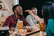 © LIGHTFIELD STUDIOS - back view of young woman feeding african american man with fried onion ring while celebrating octoberfest in pub