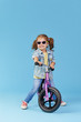© oes - Little girl learns to keep balance while riding a bicycle. Stylish child in sunglasses  isolated on blue background