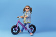 © oes - Little girl learns to keep balance while riding a bicycle. Stylish child in sunglasses  isolated on blue background