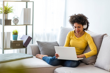 Wall Mural - Smiling young woman with headphones using laptop in livingroom. Female studying at home. Young woman wearing headphones listens online web free audio course at home