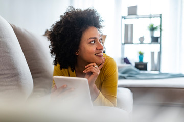 Wall Mural - Social media time. Staying in touch with her tablet. Cheerful woman sitting on couch using tablet pc at home in the living room. Smiling young woman with tablet on the sofa