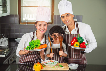 Wall Mural -  Asian woman young mother and father with son boy cooking salad food with vegetable holding tomatoes and carrots, bell peppers on plate for happy family cook food enjoyment lifestyle kitchen in home