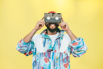 Wall Mural - Stylish young hipster man with beard in red hat, sunglasses and a retro jacket of 90s with VHS cassette on yellow background.