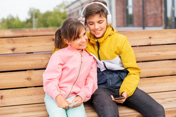 Sticker - childhood, technology and people concept - happy children or brother and sister with headphones and smartphones sitting on wooden street bench outdoors and listening to music