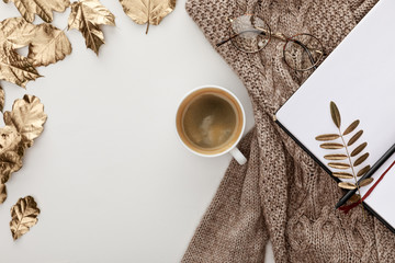 Wall Mural - top view of knitted brown sweater, glasses, coffee, blank notebook and golden foliage on white background