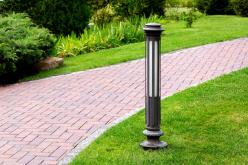 Wall Mural - ground iron lantern in a park on a green lawn with green bushes and flowerbed at a pedestrian footpath of red tile, nobody.