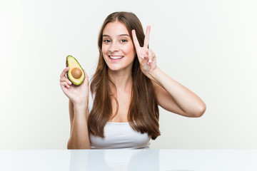 Young caucasian woman holding an avocado showing victory sign and smiling broadly.