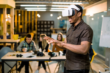 African young man wearing virtual reality glasses in modern interior design coworking studio. Smartphone using with VR goggles headset. Team of young people working at the table on the background