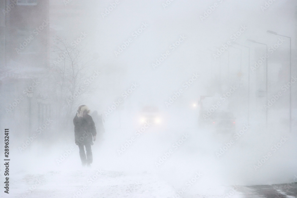 Snowstorm in the city. A man during a blizzard is walking along the street. Cars on a snowy road. Strong wind and snowfall. Arctic climate. Extreme North. Anadyr, Chukotka, Siberia, Far East Russia. - obrazy, fototapety, plakaty 