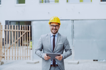 Handsome caucasian happy architect in gray suit and with yellow helmet on head holding tablet while standing on construction site.
