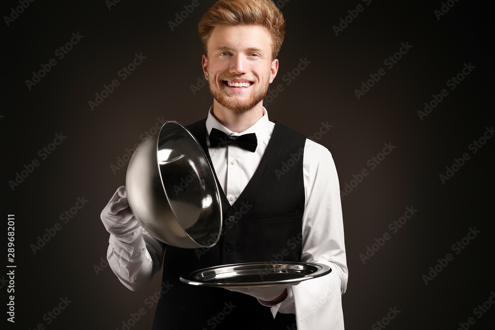 Handsome waiter with empty tray and cloche on dark background