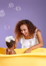 Young African-American Mother Washing Her Baby In Bathtub Against Color Background