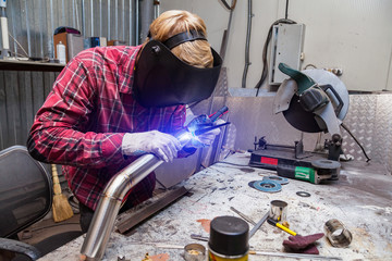 Canvas Print - Young guy welder in a checkered red shirt welds a stainless steel pipe using agronomic welding to protect his eyes with a mask in an iron workshop. Modern welding methods.