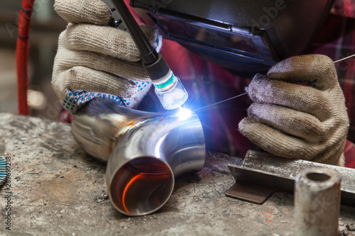 Young guy welder in a checkered red shirt welds a stainless steel pipe using agronomic welding to protect his eyes with a mask in an iron workshop. Modern welding methods.