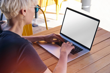  Woman with short hair working on laptop, screen space for design layout. Sitting outside in modern cafe, sunny day, having rest.