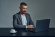 © nazarovsergey - Bearded businessman with stylish mustache, dressed in a classic suit is working at laptop while sitting at table in office, gray background.