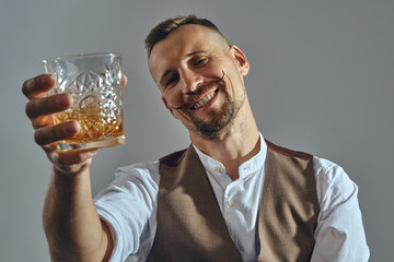 Wall Mural - Man with stylish mustache, dressed in classic brown vest, white shirt is sitting at the table, enjoying whiskey. Grey background, close-up shot.