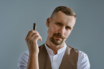 Wall Mural - Man with a stylish mustache, dressed in brown vest and white shirt is posing sitting on a table, enjoying cigar. Grey background, close-up.