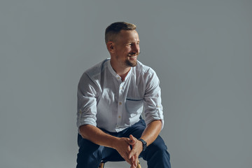 Wall Mural - Man with stylish mustache, in classic white shirt and blue trousers is posing sitting on chair. Grey background, close-up shot.