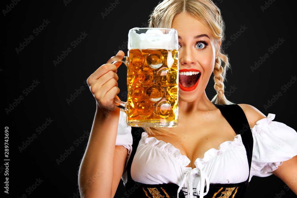 Smiling young sexy oktoberfest girl waitress, wearing a traditional Bavarian or german dirndl, serving big beer mug with drink isolated on black background. - obrazy, fototapety, plakaty 