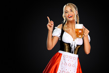 Smiling Sexy Oktoberfest Girl Waitress, Wearing A Traditional Bavarian Or German Dirndl, Serving Big Beer Mug With Drink Isolated On Black Background. Woman Pointing To Looking Left.