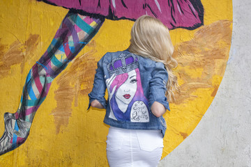 The blonde poses in a painted denim jacket on the background of the wall. On the street.