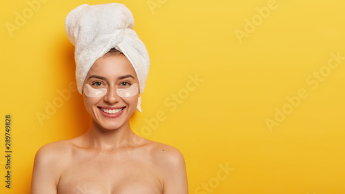 Fotomural Studio shot of beautiful young woman with healthy delicate fresh skin under eyes, wears towel on head, applies patches for reducing fine lines, stands naked against yellow background