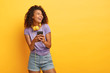 Photo of smiling teenage girl with Afro haircut, uses smartphone for listening music in playlist, wears headphones, looks positively aside, dressed in everyday summer clothes copy space on yellow wall