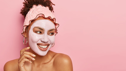 Wall Mural - Smiling charming young woman cares about skin, applies clay mask, nourishes complexion with moisturizing cream, wears pink headband, round earrings, stands bare shoulders, looks happily aside