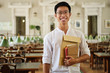 Young smiling asian student in eyeglasses with notepads joyfully looking in camera in library of university