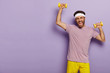 Horizontal shot of motivated sportsman trains muscles, raises yellow dumbells, wears headband, casual outfit, being active, poses over purple studio wall, wants to have strong biceps. Sporty lifestyle