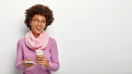 Positive Afro American looks with carefree expression, holds modern call phone, surfs net, sends text messages, enjoys drinking aromatic coffee, dressed in purple outfit, wears optical glasses