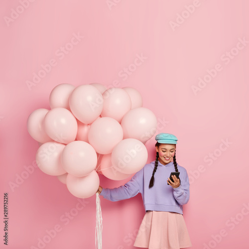 People, holiday, celebration, technology concept. Positive brunette girl types messages on mobile phone, surfs internet, carries helium balloons, poses against pink background with empty space above