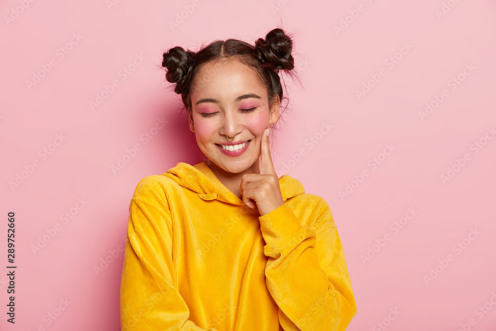 Positive lovely woman with pinup makeup, keeps index finger on cheeks, has eyes closed, dreams about something pleasant, wears yellow velvet hoody, models against pink background. Natural beauty - obrazy, fototapety, plakaty 
