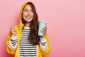 Wall Mural - Positive woman with long dark straight hair, raises clenched fist, holds takeaway coffee, dressed in striped jumper, yellow raincoat, enjoys hot drink during cold rainy autumn day, feels calm, relaxed