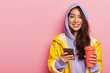 Delighted attractive Asian woman wears casual clothes, raincoat, sends message via cell phone, drinks aromatic beverage from thermos, being in good mood, always in touch. Season, leisure, lifestyle