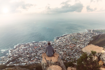 Wall Mural - Young man sitting on the edge at the top of Lion's head mountain in Cape Town with a beautiful evening city view