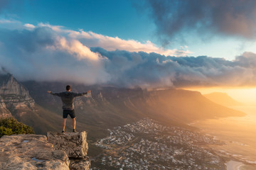 Wall Mural - Young man standing on the edge at the top of Lion's head mountain in Cape Town with a beautiful sunset view