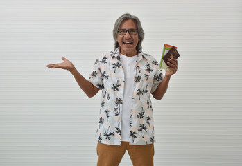 Happy senior traveler asian man wearing glasses and summer shirt holding travel map and Thailand passport, shows outstretched hand with open palm standing over white wall background, Business summer h