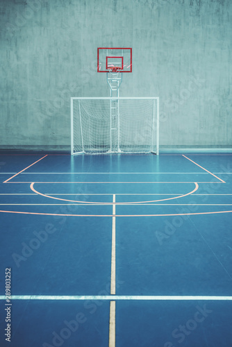 Front view of the court in the gym hall; indoors modern contemporary office stadium with a basketball basket and hoop, football goal, colored marking on the floor, a concrete wall in the background