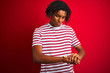 © Krakenimages.com - Young afro man with dreadlocks wearing striped t-shirt standing over isolated red background Checking the time on wrist watch, relaxed and confident