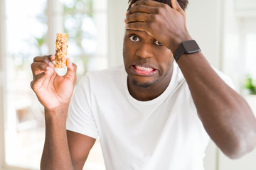 Poster - African american man eating energetic cereals bar stressed with hand on head, shocked with shame and surprise face, angry and frustrated. Fear and upset for mistake.