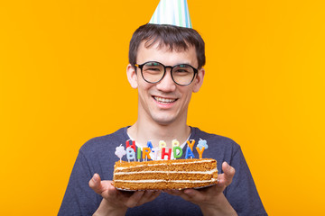 Wall Mural - Funny positive guy in glasses holds in his hands a homemade cake with the inscription happy birthday posing on a yellow background. Concept of holidays and anniversaries.