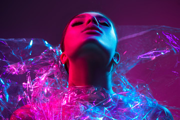 High Fashion model girl in colorful bright neon lights posing in studio through transparent film. Portrait of beautiful sexy woman in UV. Art design colorful make up. On colourful vivid background