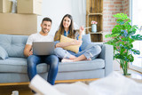 Fototapeta Młodzieżowe - Young beautiful couple relaxing sitting on the sofa of new house using computer laptop very happy pointing with hand and finger to the side