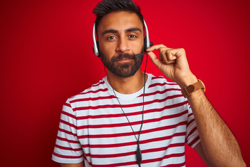 Young indian call center agent man using headset over isolated red background with a confident expression on smart face thinking serious