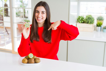 Poster - Beautiful young woman eating half fresh green kiwi with surprise face pointing finger to himself