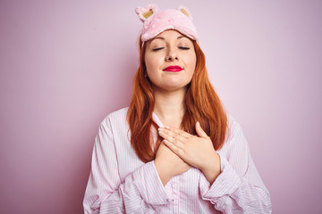 Poster - Young beautiful redhead woman wearing pajama over pink isolated background smiling with hands on chest with closed eyes and grateful gesture on face. Health concept.